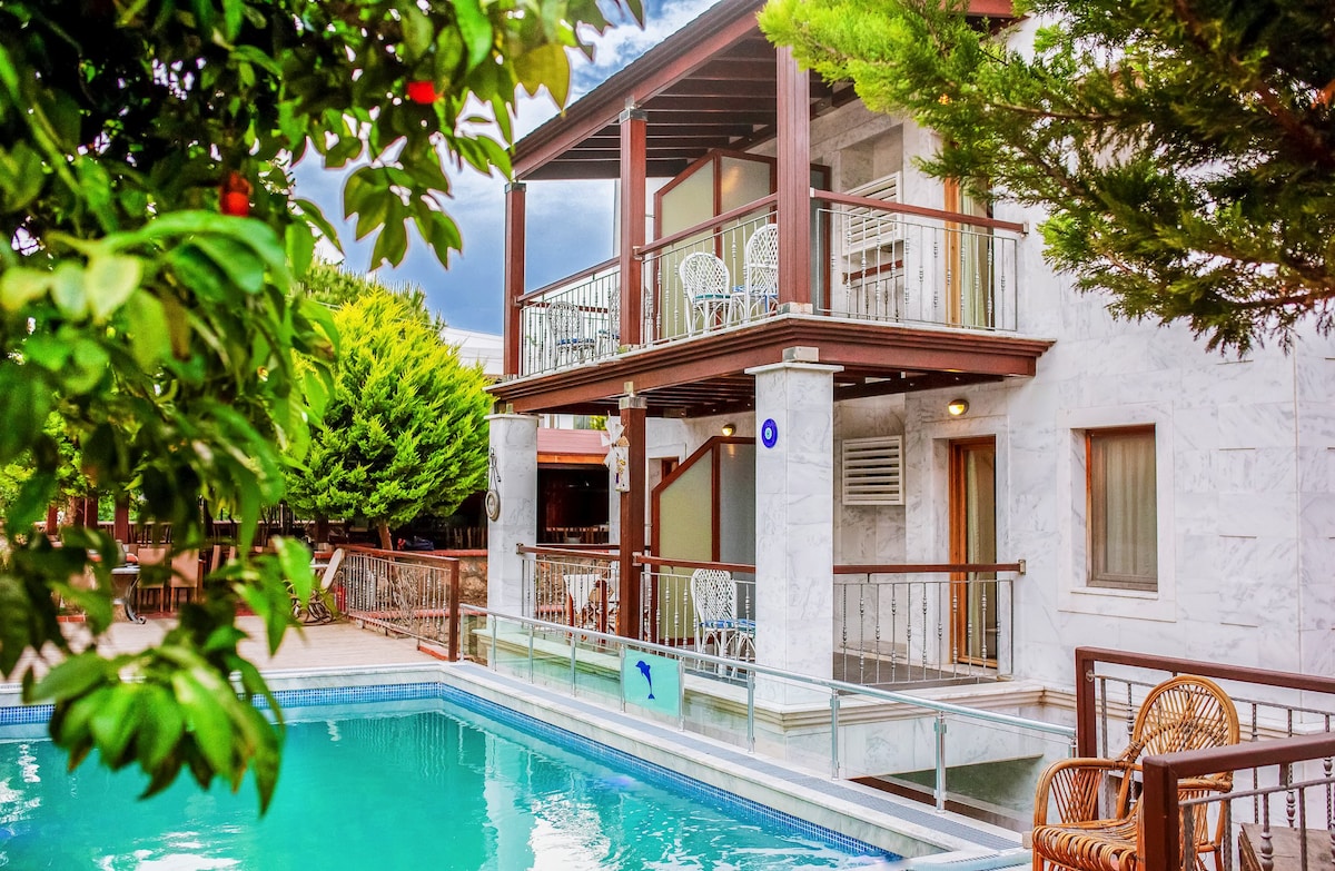 A white stone building showcases a modern architectural style with multiple balconies overlooking a clear pool surrounded by greenery. Comfortable wicker chairs are visible on the balcony, inviting relaxation. The trees provide shade, enhancing the tranquil atmosphere of the outdoor space.