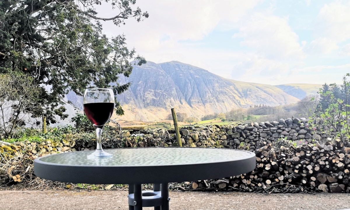 A glass of red wine is placed on a round, glass-top table, with a panoramic view of rolling hills and a mountainside in the background. Stone walls and greenery are visible, enhancing the natural scenery.