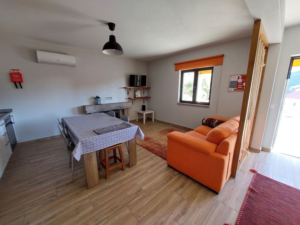 An open living area features a cozy orange sofa and a dining table with wooden chairs. A kitchenette is visible in the background, complemented by a light-colored countertop. Natural light enters through a window, enhancing the simple yet functional layout.
