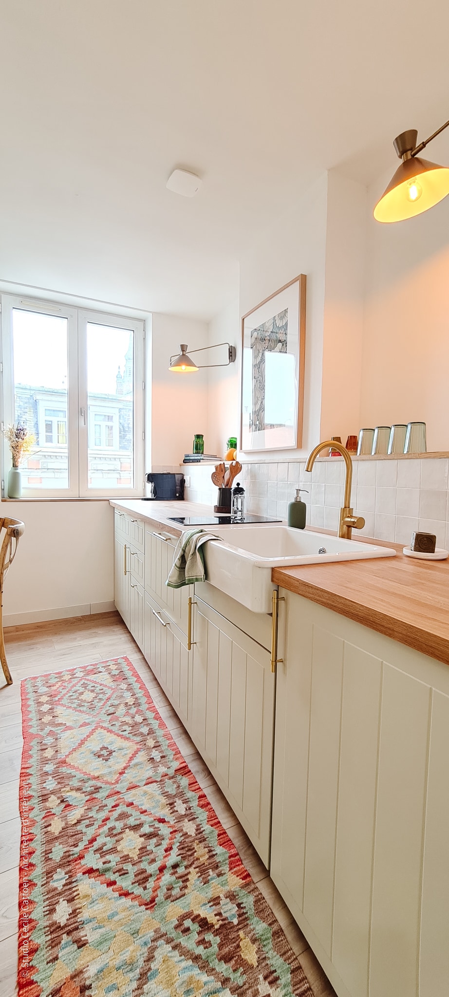 The kitchen features contemporary cabinetry and light wood countertops, complemented by a farmhouse sink. Natural light enters through a large window, illuminating the space. A colorful area rug adds warmth, while simple decor accents enhance the modern design.