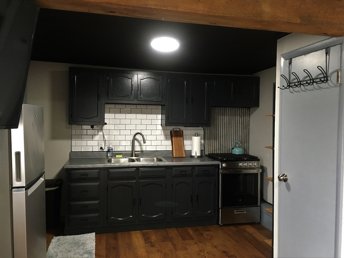 The kitchen area features dark cabinetry contrasted by white subway tiles and a modern stainless steel stove. A single basin sink is positioned beneath a circular ceiling light, with space for cooking equipment and utensils. The flooring is hardwood, complementing the overall aesthetic.