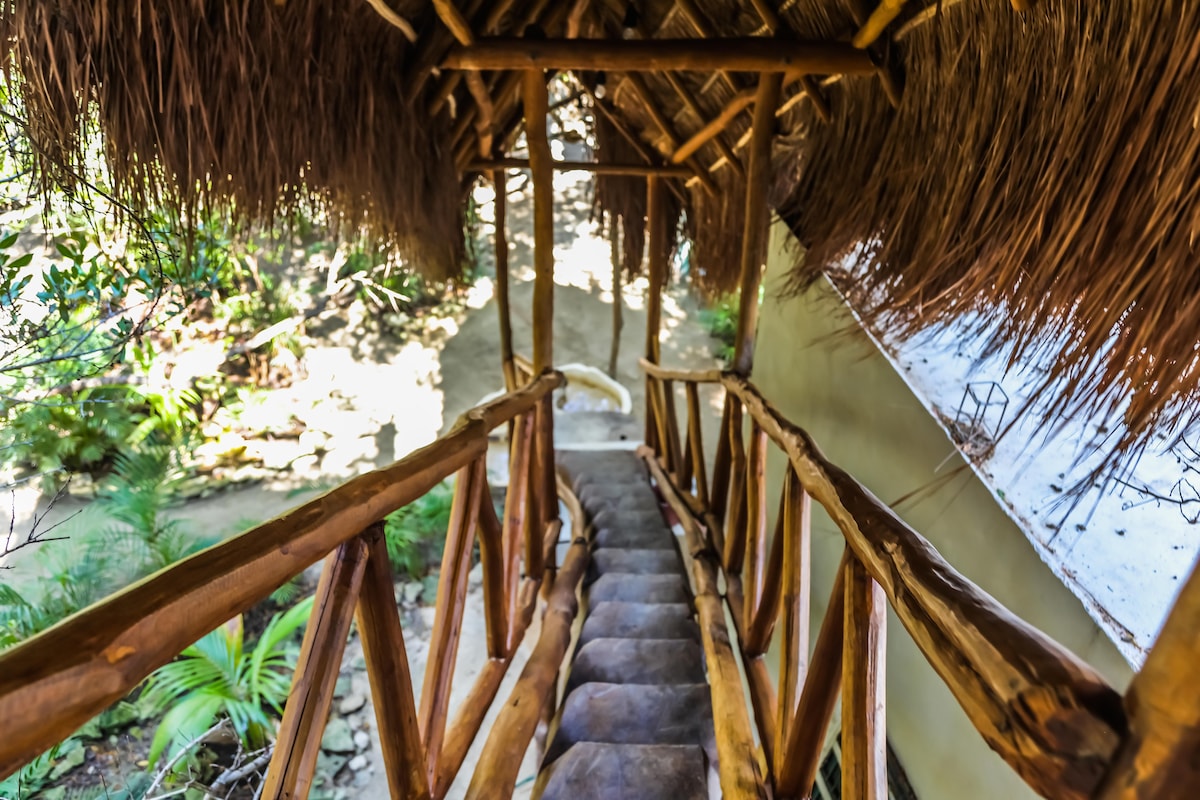 A wooden staircase is visible, leading downwards through a covered pathway adorned with a thatched roof. Surrounding greenery is present, adding a natural touch to the environment.