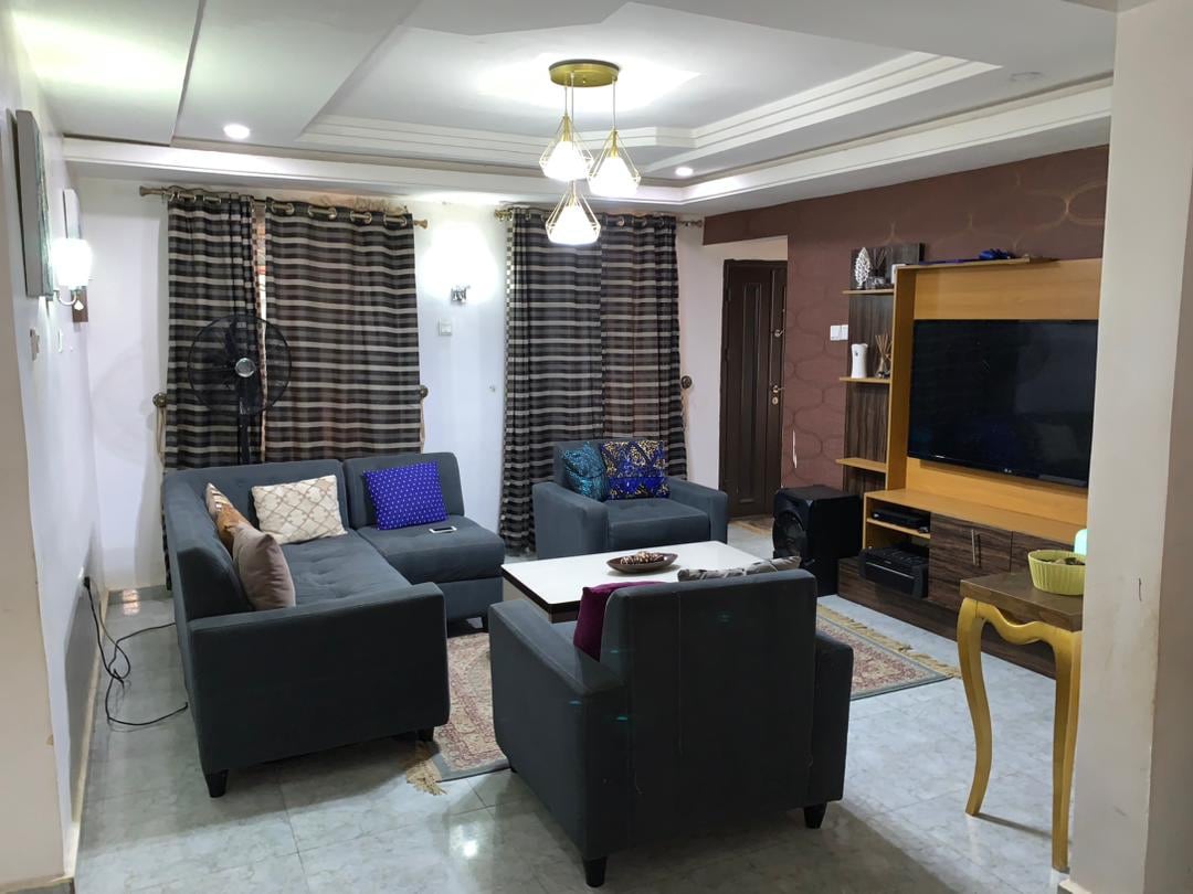 A living area is presented with a dark upholstered sectional sofa and scattered cushions in various colors. A coffee table occupies the center, while a television is positioned on a wooden unit against a textured wall. Natural light is filtered through the dark-striped curtains.