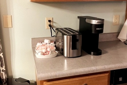 A countertop is displayed with a Keurig coffee maker and a silver toaster. Nearby, a white dish holds coffee creamers, providing convenience for morning routines. The backdrop consists of wooden cabinetry and a light-colored wall, enhancing the functional kitchen area.