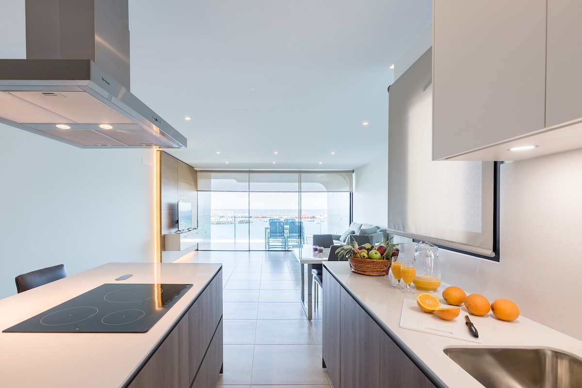 A modern kitchen is displayed with a central island featuring a cooktop and ample counter space. Fresh fruit is arranged on the counter. Large glass doors provide a view of the outdoor terrace and ocean, allowing natural light to fill the open-concept living area.