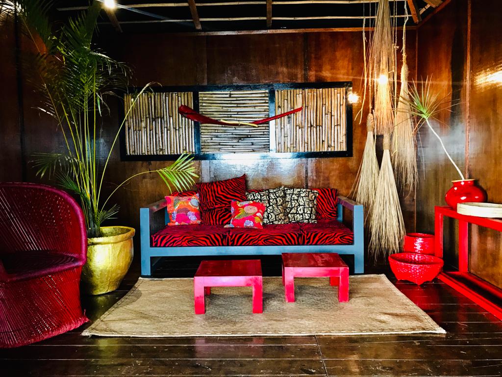A vibrant living area is depicted, featuring a comfortable seating arrangement with colorful cushions on a red-and-gray sofa. Decor elements include a woven wall art piece, potted plants, and decorative tables, creating an inviting space that reflects unique bohemian style.