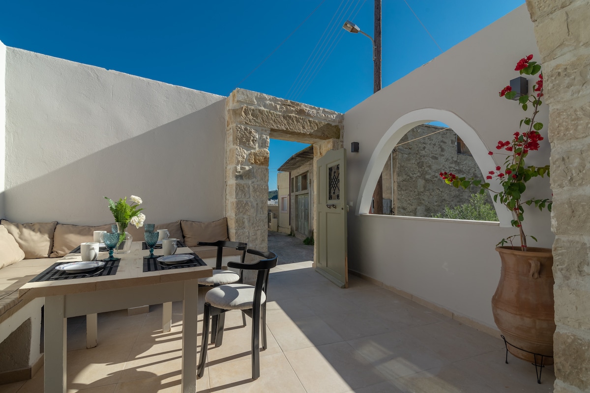 An outdoor dining area features a wooden table set with dishes and beverages, surrounded by soft seating. A stone archway leads to a pathway, while a potted plant adds a splash of color. The clear blue sky enhances the tranquility of the space.