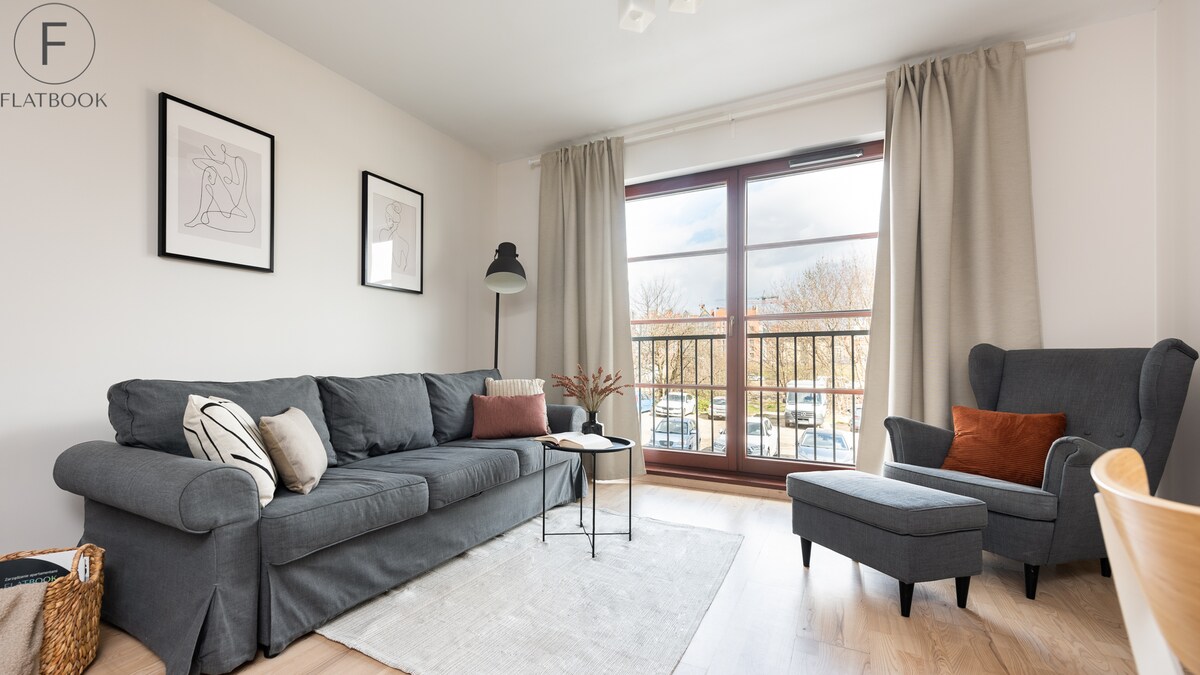 A modern living area is presented with a comfortable gray sofa and an accent chair. Large windows allow natural light to fill the space, with neutral curtains framing the view. A small side table holds decorative items, adding a touch of warmth to the ambiance.
