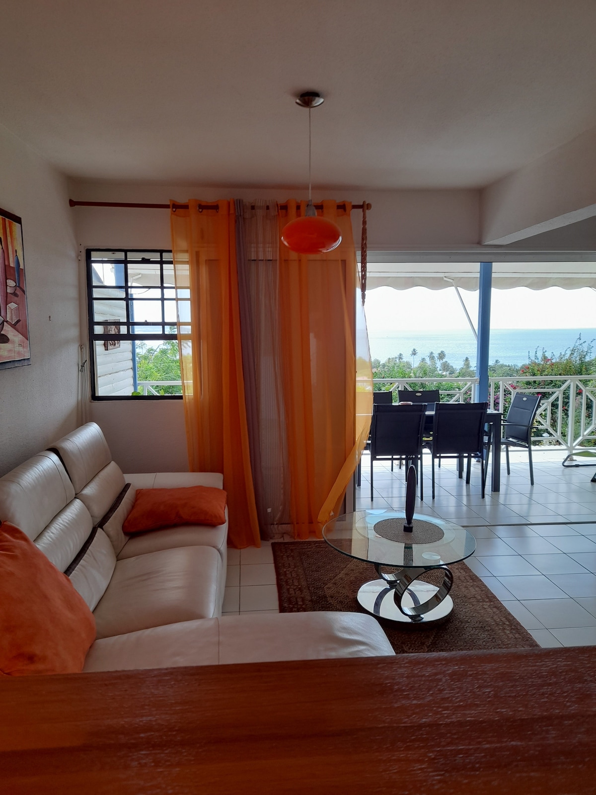 A cozy living area is highlighted by bright orange curtains, which frame a large window offering views of the outside. A light-colored sofa with orange cushions is arranged around a circular glass coffee table. Dining chairs and a table are visible on the terrace.