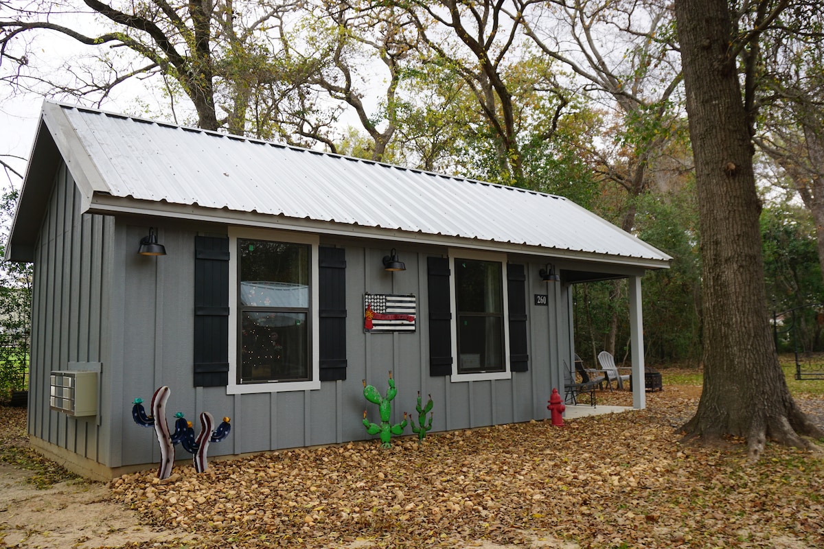A tiny house with a gray exterior and a metal roof is nestled among autumn leaves. Two windows are framed by black shutters, with decorative elements including a firefighter's flag and cacti. A small porch features a couple of chairs, creating a welcoming outdoor space.