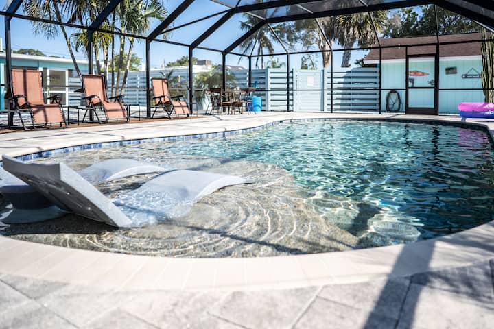 A newly heated pool is showcased within a screened enclosure, surrounded by lounge chairs. Sunlight reflects on the clear water, while a small platform extends into the pool, creating a serene atmosphere for relaxation.