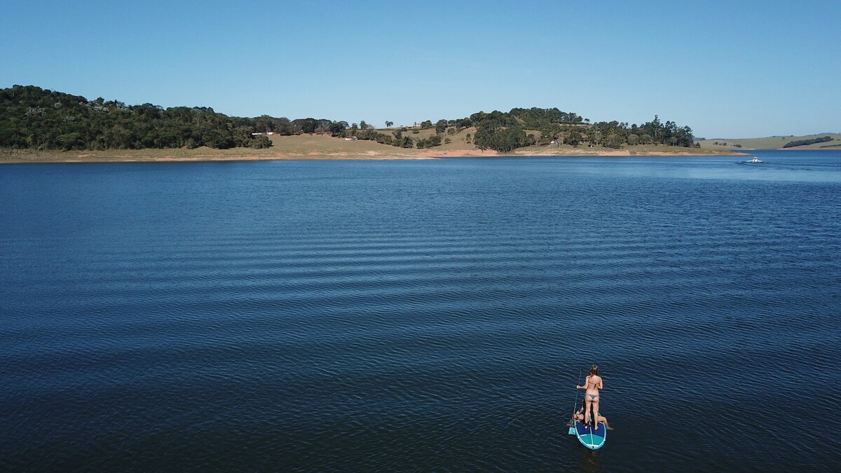 A clear view of tranquil water surfaces a large reservoir bordered by green hills. A person is seen standing on a paddleboard, creating gentle ripples across the lake, which reflects the expansive blue sky above. Nature's serenity is highlighted in the setting.
