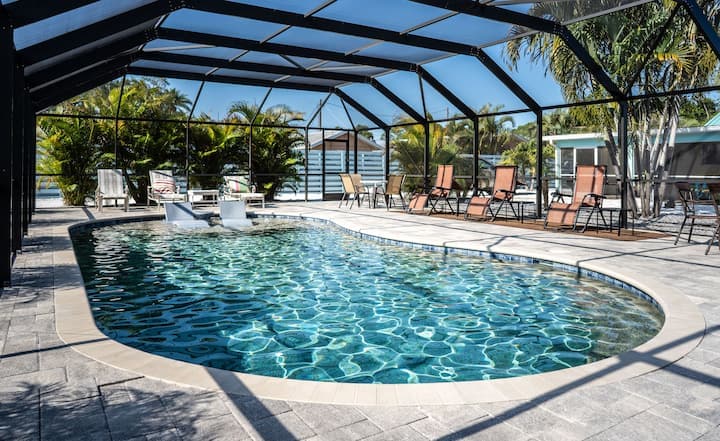 A heated saltwater pool is enclosed within a screened area, featuring lounge chairs positioned both along the poolside and on a wooden deck. Lush greenery is visible beyond the enclosure, offering a sense of privacy and tranquility.