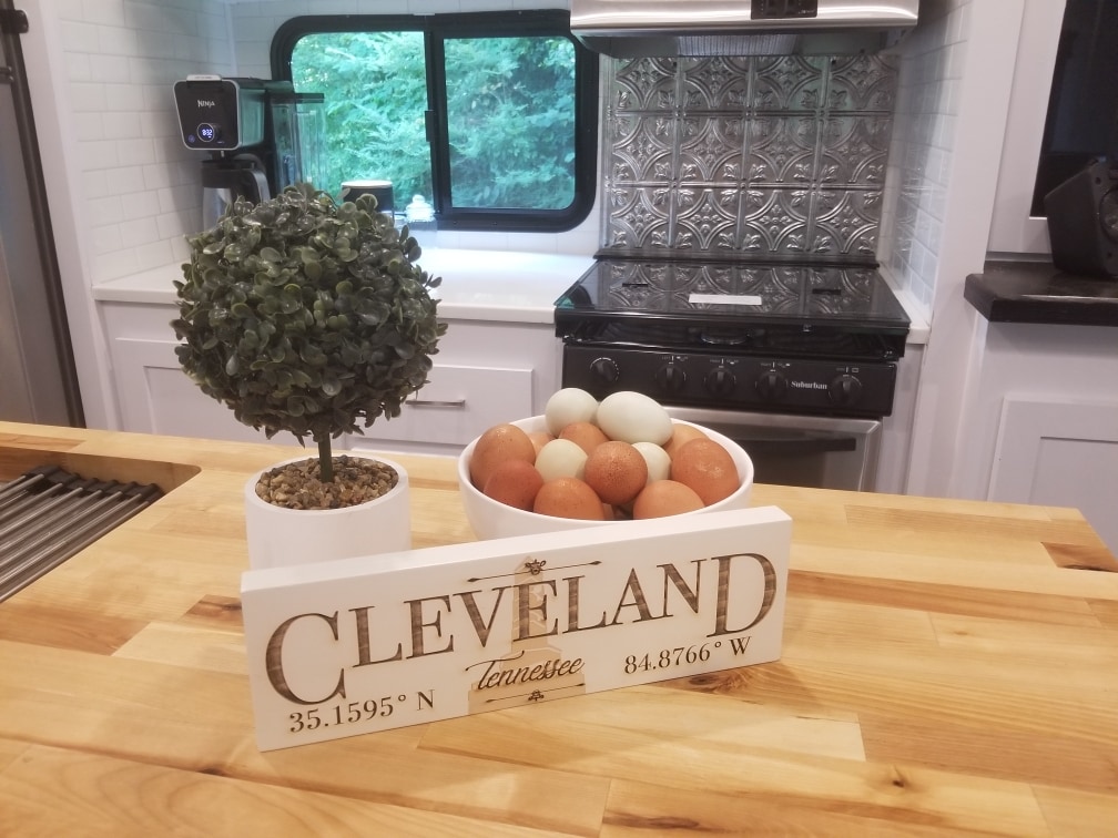 A kitchen countertop features a decorative topiary and a white dish filled with eggs, providing a natural touch to the space. Behind, the stainless-steel stove and tiled backsplash highlight a modern aesthetic, complemented by a glimpse of greenery visible through the window.