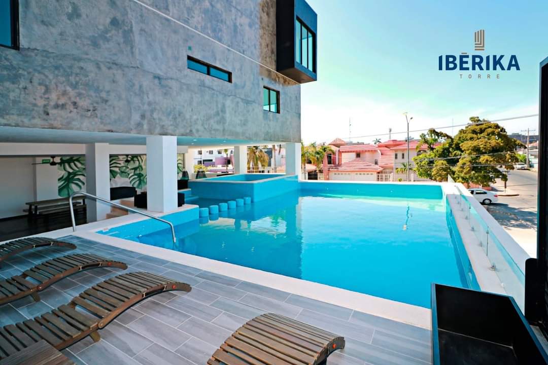 A modern swimming pool is shown, featuring clear blue water and surrounded by wooden lounge chairs. The pool area is part of a sleek building and offers a view of the surrounding neighborhood, with ample natural light illuminating the space.