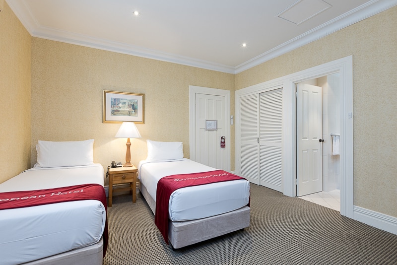 A well-lit guest room features two spacious beds, each dressed with crisp white linens and red accents. A bedside table with a lamp is positioned between the beds. A closet and bathroom door are visible along the wall, contributing to the room's functional layout.