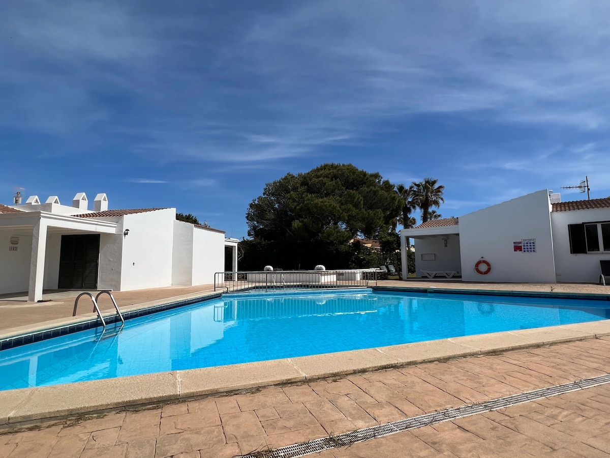 A communal swimming pool is surrounded by a spacious tiled area. The clear blue water reflects the sky, while a lush green tree offers shade nearby. White buildings are positioned on either side of the pool, contributing to a tranquil environment.