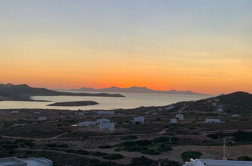 A breathtaking sunset casts warm hues over the landscape, highlighting the coastline and distant islands. The view captures a serene bay with gentle waves and scattered white buildings nestled among green vegetation, creating a peaceful atmosphere as day transitions to night.