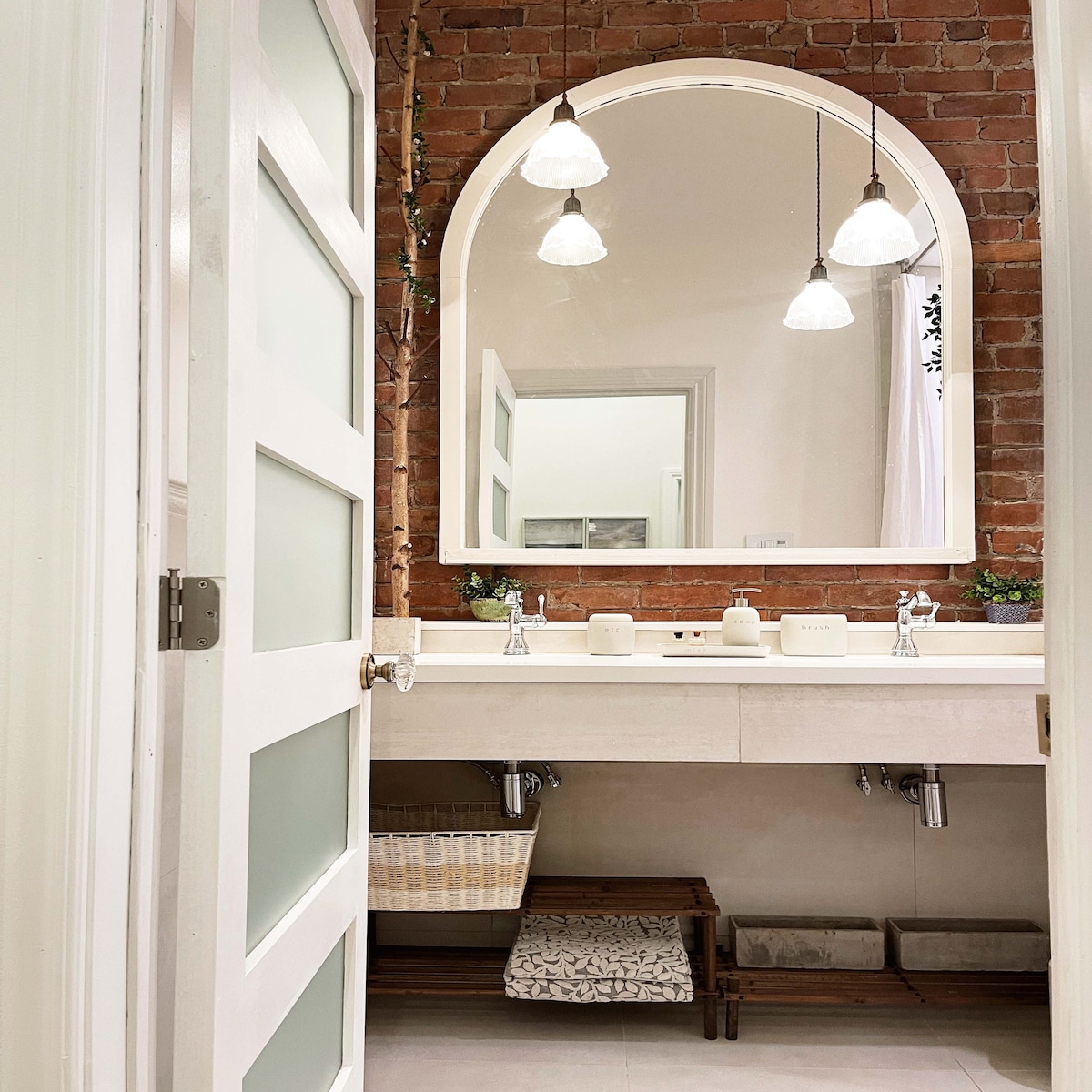 A modern bathroom features a wall of exposed brick, providing a warm contrast to the light-colored vanity with dual sinks. Above the sinks, arched mirrors are illuminated by three pendant lights. Storage options include an open shelf and a woven basket.
