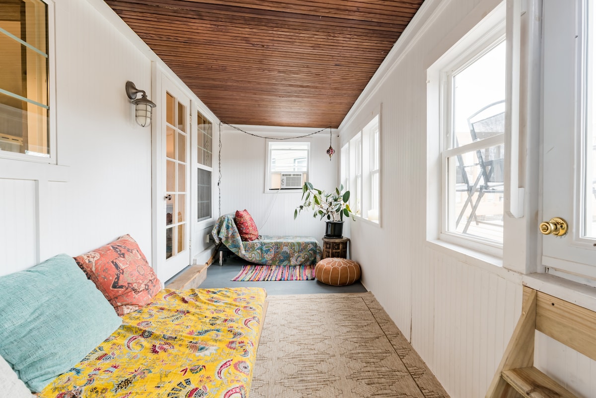 A bright and airy sunroom features a vibrant area rug and two comfortable seating spaces. Natural light filters through multiple windows, accenting the decorative throw pillows and unique wall art. A small indoor plant adds a touch of greenery to the inviting setting.