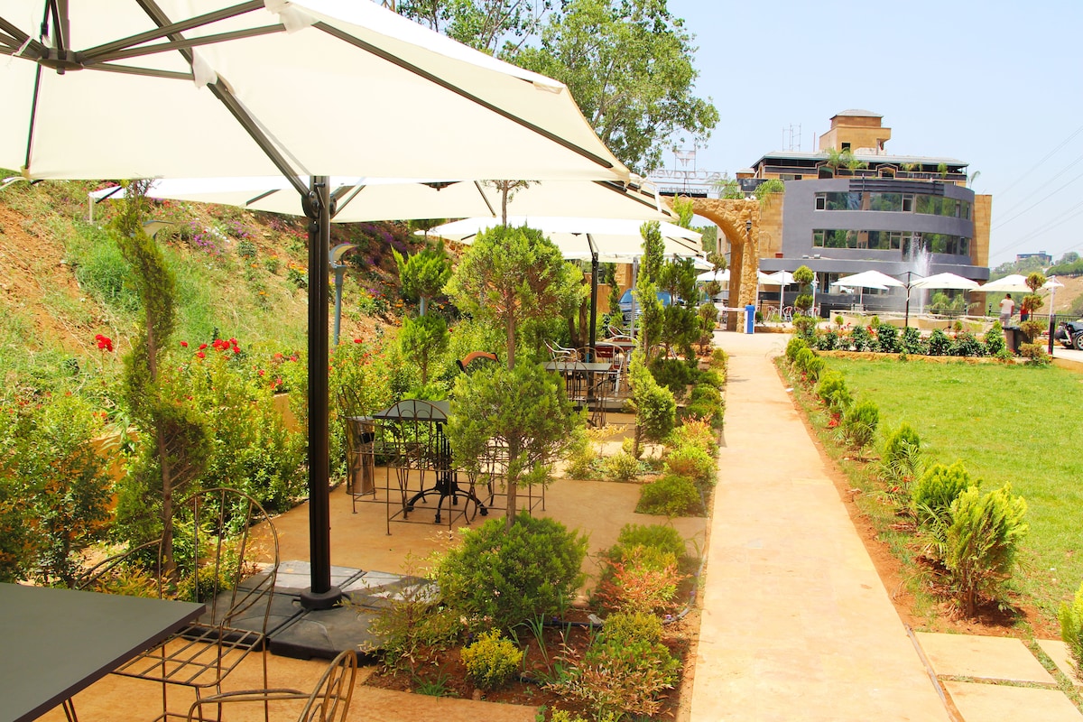 Outdoor seating areas are provided under large umbrellas, surrounded by neatly trimmed bushes and flower beds. A stone pathway leads through the landscaped garden, with a modern building visible in the background, offering a relaxed atmosphere for dining or socializing.