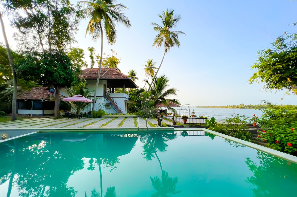 The image captures a tranquil swimming pool reflecting the surrounding palm trees and the clear sky. A two-story cottage is visible in the background, nestled among lush greenery, with outdoor seating areas positioned near the lake. The pristine waters of Vembanad Lake are seen beyond the property.