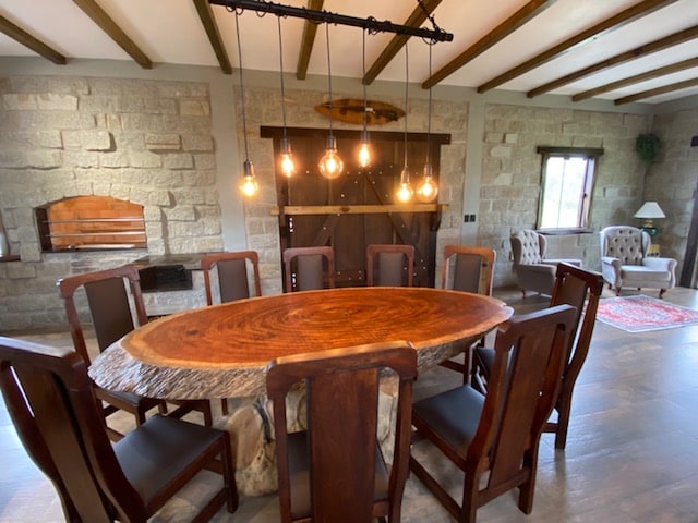 A large dining area is featured with an oval wooden table surrounded by ten dark wooden chairs. Industrial-style pendant lights hang above the table, providing illumination. Stone walls and a barn-style door emphasize the rustic design, while a cozy seating area is visible in the background.