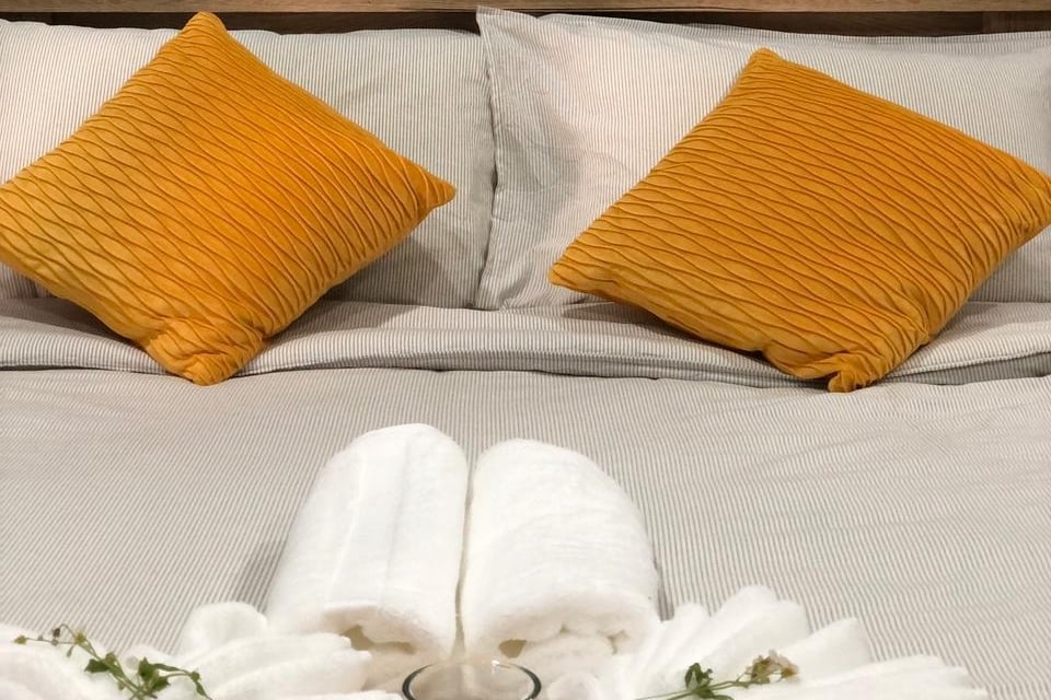 A spacious bed is dressed in soft grey linens, flanked by two vibrant orange pillows. White towels are elegantly arranged in a heart shape at the foot of the bed, complemented by a small decorative candle.