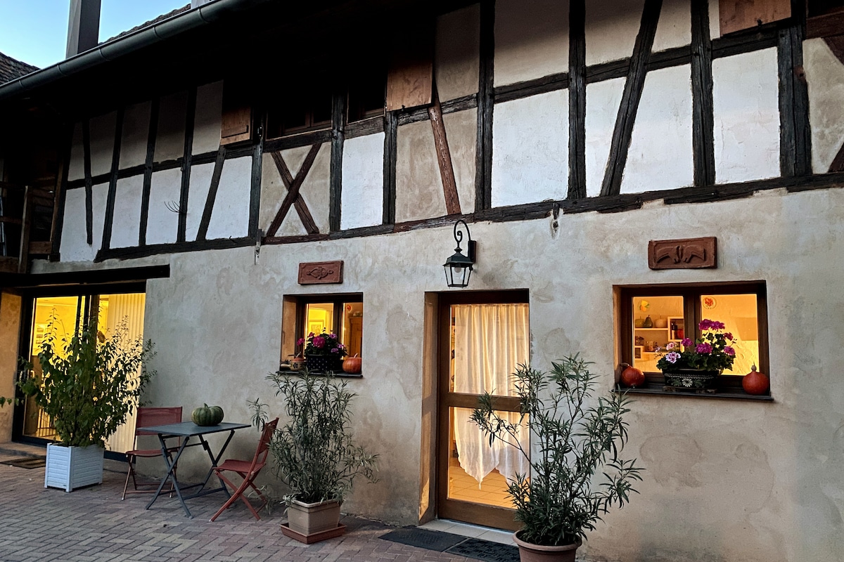 The exterior of a renovated traditional Alsatian farmhouse is displayed, featuring a textured wall with exposed wooden beams. Two window boxes filled with vibrant flowers add colour, while a small table and chairs are positioned outside, offering a space for relaxation.