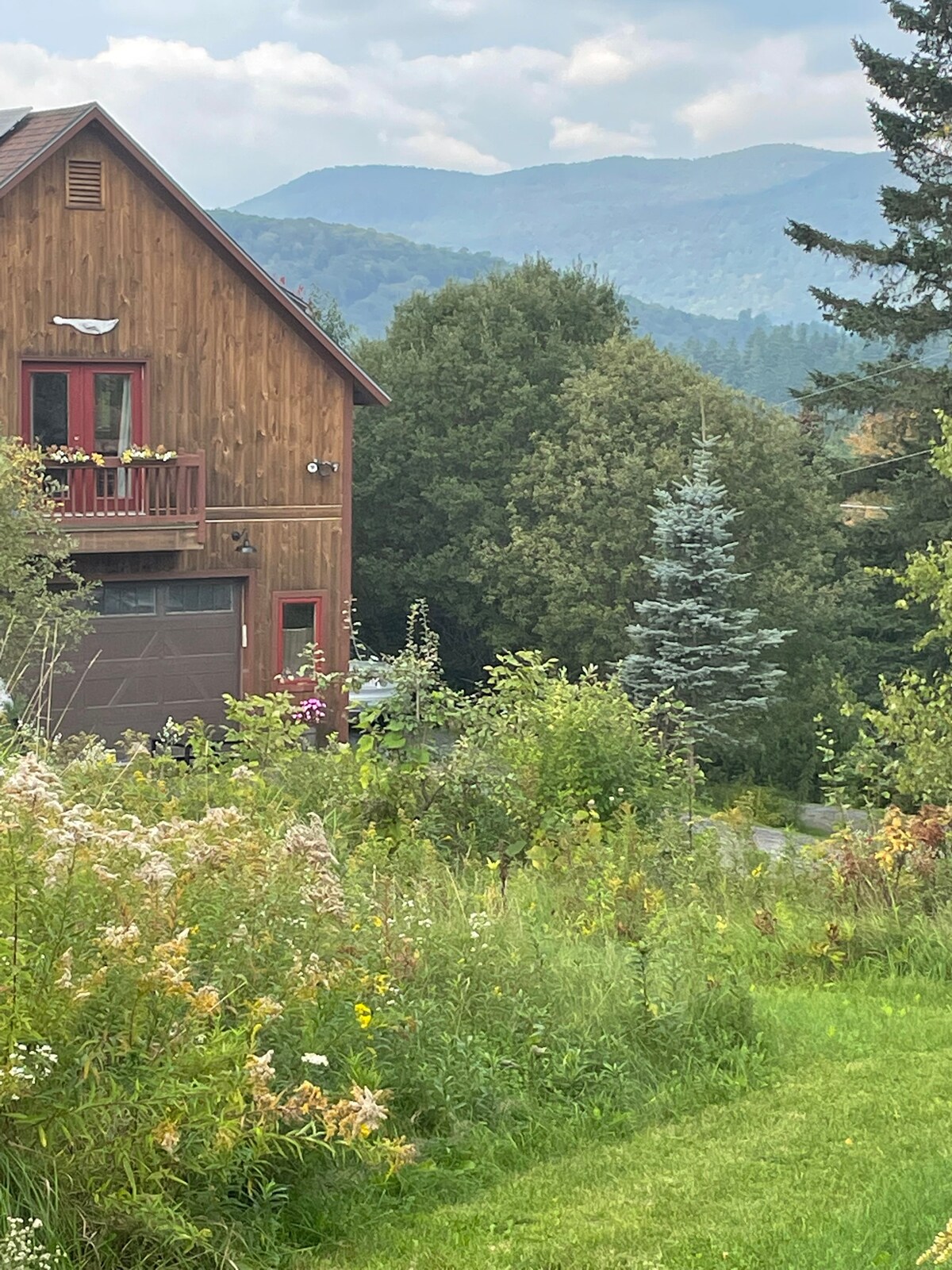 A charming wooden house is framed by vibrant greenery and wildflowers. Majestic mountains rise in the background, partially shrouded by soft clouds. The mixture of trees and foliage creates a serene and natural setting.