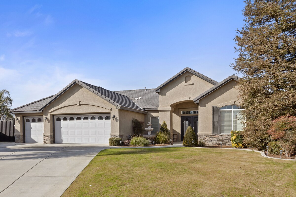 A single-story home with a neutral exterior presents a welcoming entrance highlighted by a rounded archway. The spacious driveway accommodates multiple vehicles and is flanked by well-maintained landscaping, including shrubs and small trees, leading to the entrance.