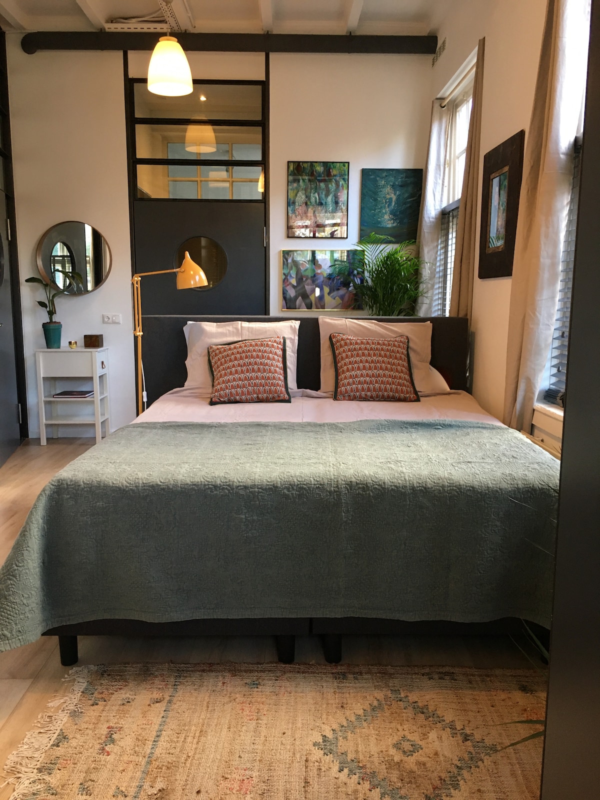 A well-appointed sleeping area features a spacious bed adorned with an aqua-colored blanket and patterned cushions. A round mirror and a potted plant are positioned nearby. Colorful artwork decorates the walls, complementing the natural light filtering through the window.