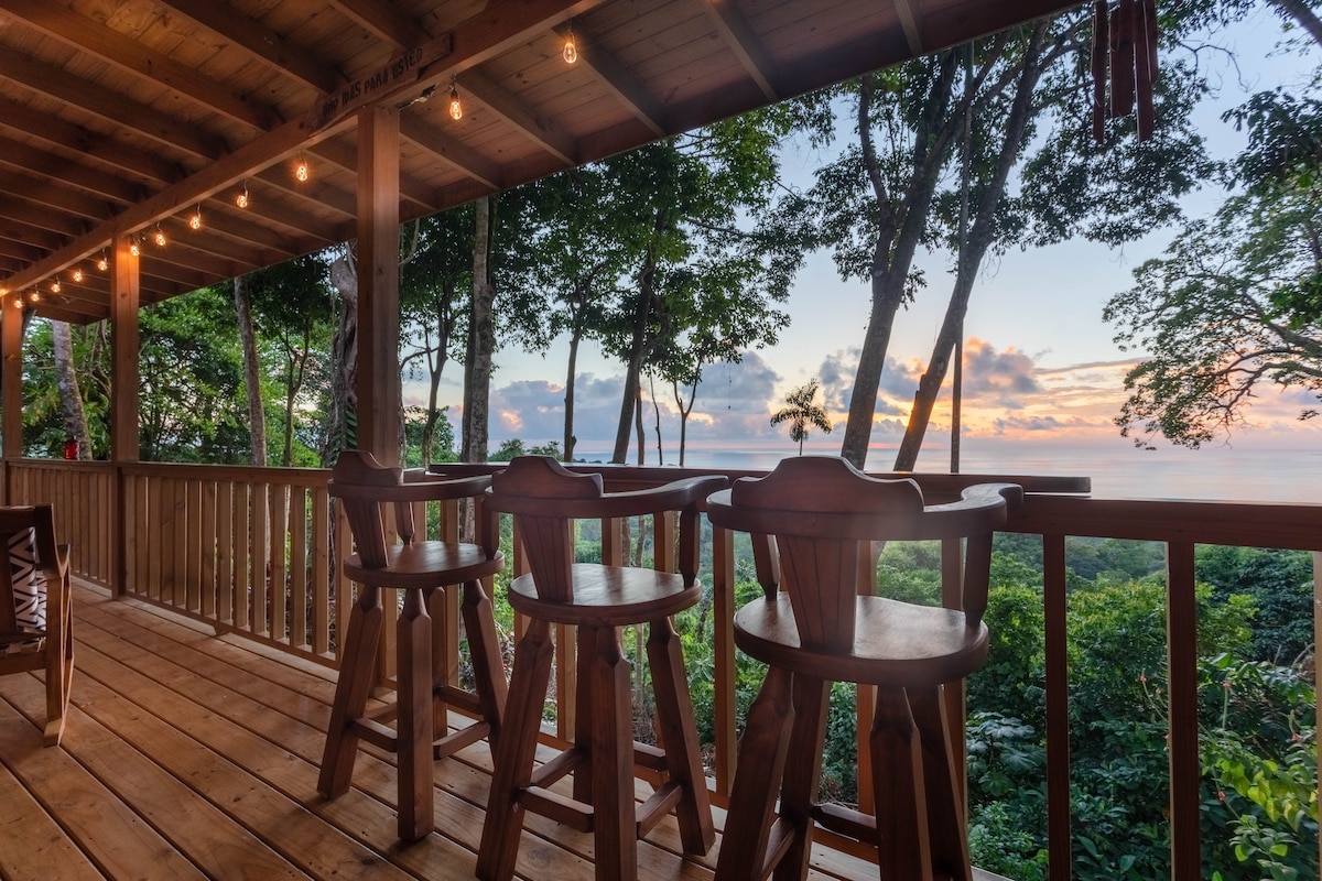A wooden patio features high stools facing the ocean, set against a backdrop of lush greenery. Soft evening light illuminates the space, while trees frame the horizon and colorful clouds hang above the distant water.