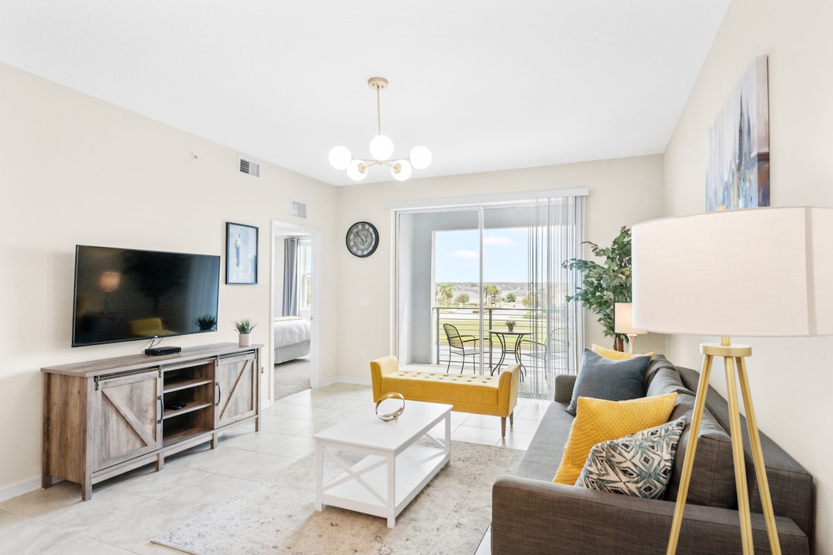 A bright living area features a comfortable gray couch adorned with patterned cushions. A stylish coffee table is positioned in front, with a yellow accent chair nearby. A large flat-screen TV is mounted on a wooden cabinet, and natural light floods the space from sliding glass doors that lead to a balcony.