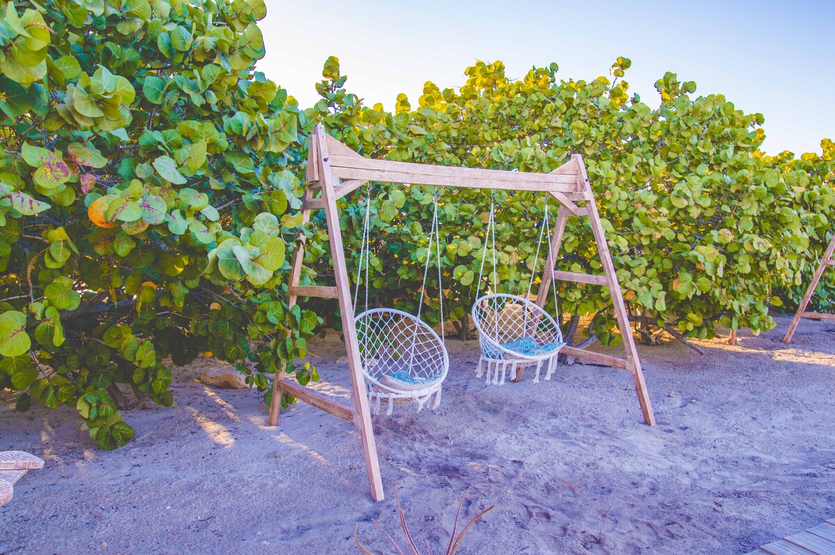 A wooden swing set is situated amidst lush greenery, featuring two basket-style swings gently hanging from a sturdy frame. The sandy ground beneath complements the natural surroundings, creating a serene space for relaxation.