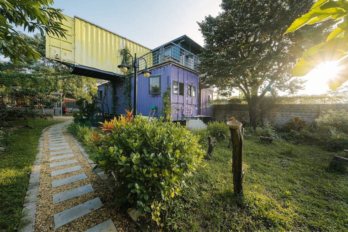 The exterior of the house features a unique design with colorful shipping containers. A stone pathway winds through a lush garden filled with greenery, leading to the entrance. The warm light of the sun filters through the trees, creating a calm and inviting atmosphere.