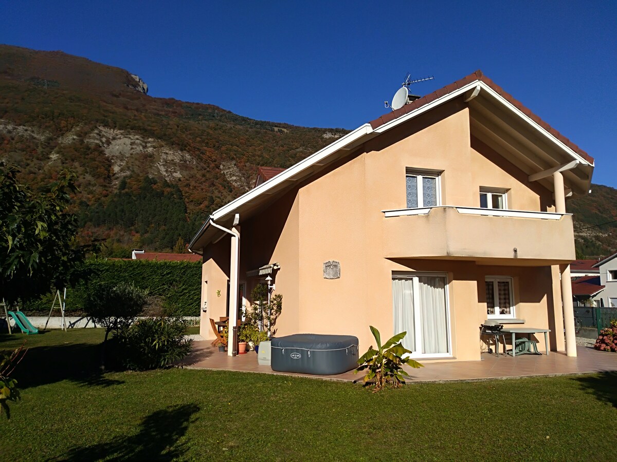 The exterior of a light-colored house is visible, showcasing a well-maintained garden area. A hot tub is placed on a patio, with a table and chairs positioned nearby. The backdrop features mountains under a clear blue sky.