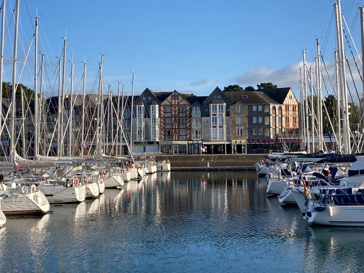 A marina scene is depicted, with several boats gently bobbing on calm waters. Colorful buildings line the waterfront, reflecting the architectural diversity. A clear blue sky and distant trees complete the serene setting, inviting exploration of the surrounding area.