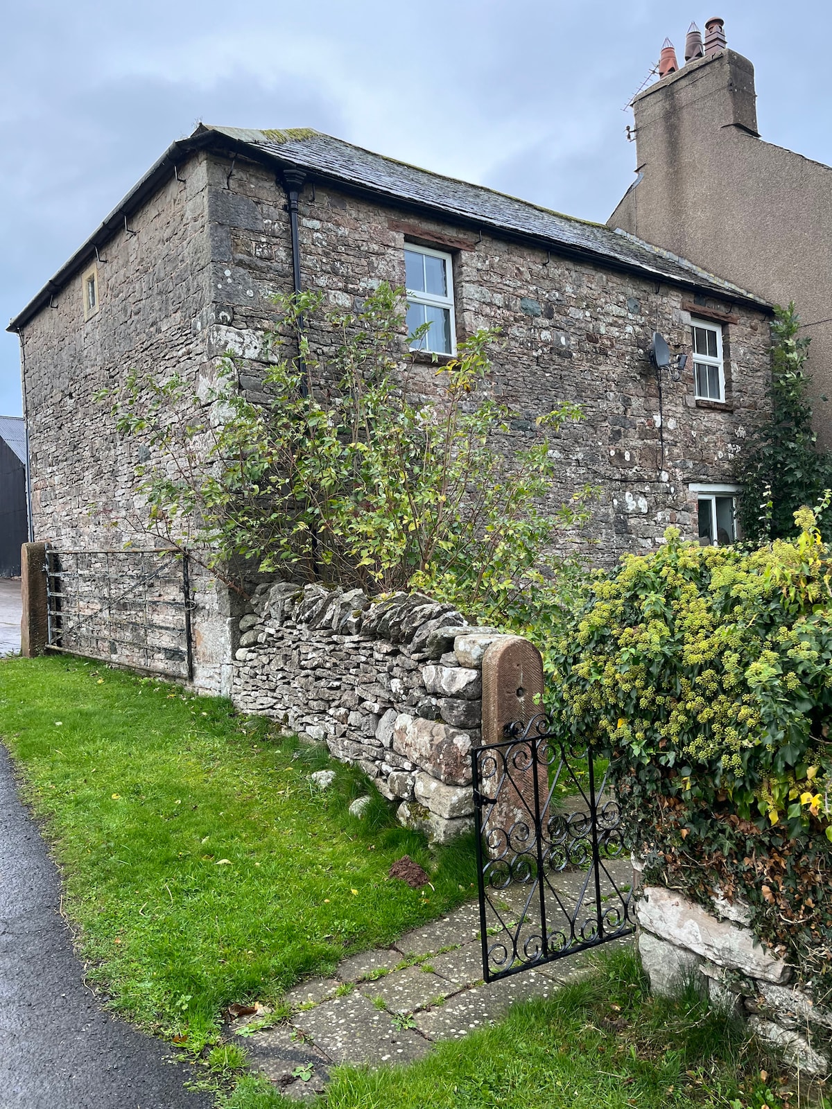 A charming stone exterior of a house is framed by green grass and a neatly manicured hedge. An iron gate leads to the pathway, while nearby foliage and a stone wall enhance the natural setting. The house features multiple windows, contributing to its inviting character.