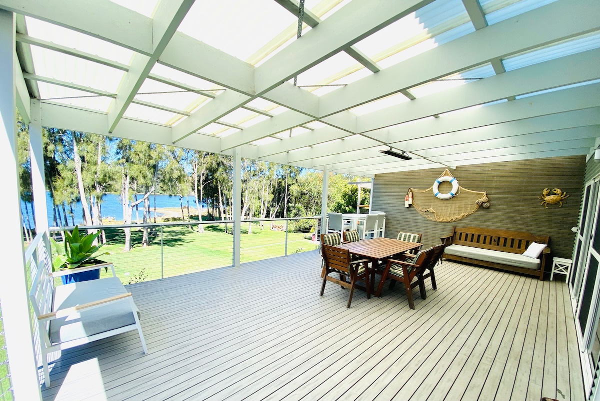A spacious covered deck features comfortable outdoor seating, a dining table, and a daybed, all set against the backdrop of lush greenery and unobstructed views of Mummuga Lake. Natural light filters through the roof, creating a bright and inviting atmosphere.
