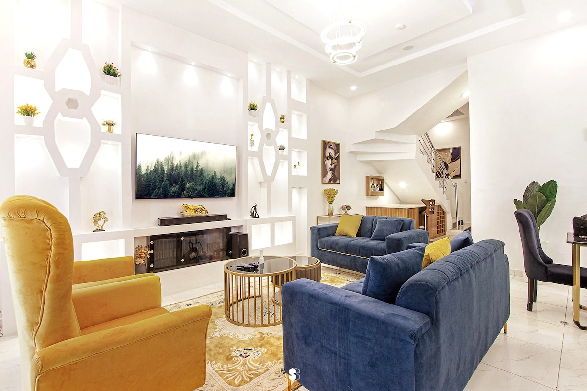 A spacious living room is showcased, featuring two plush sofas and a vibrant armchair, arranged around a central circular coffee table. A large flat-screen TV is mounted on a stylish wall adorned with decorative shelves, complemented by subtle lighting that enhances the modern design.