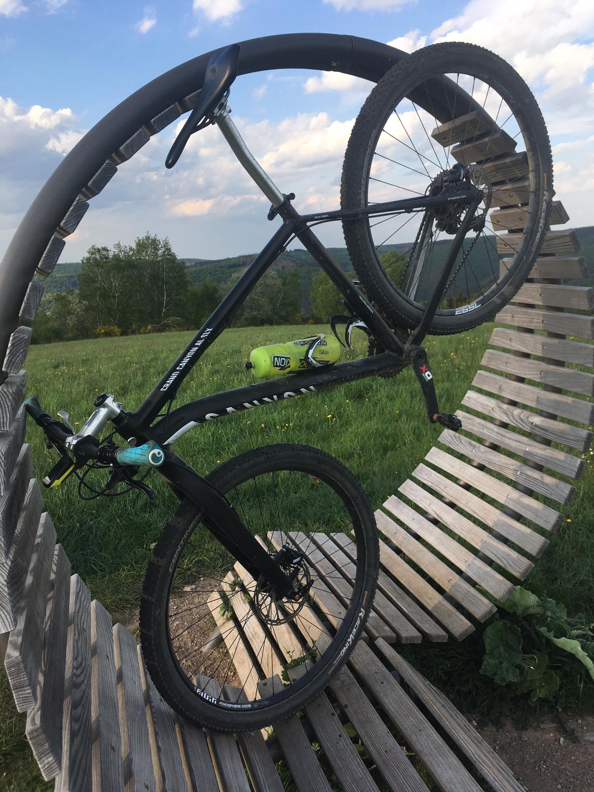 A black mountain bike is displayed on a circular wooden structure, highlighting its sturdy frame and prominent wheels. The background features green hills under a partially cloudy sky, creating a serene outdoor setting ideal for biking enthusiasts.