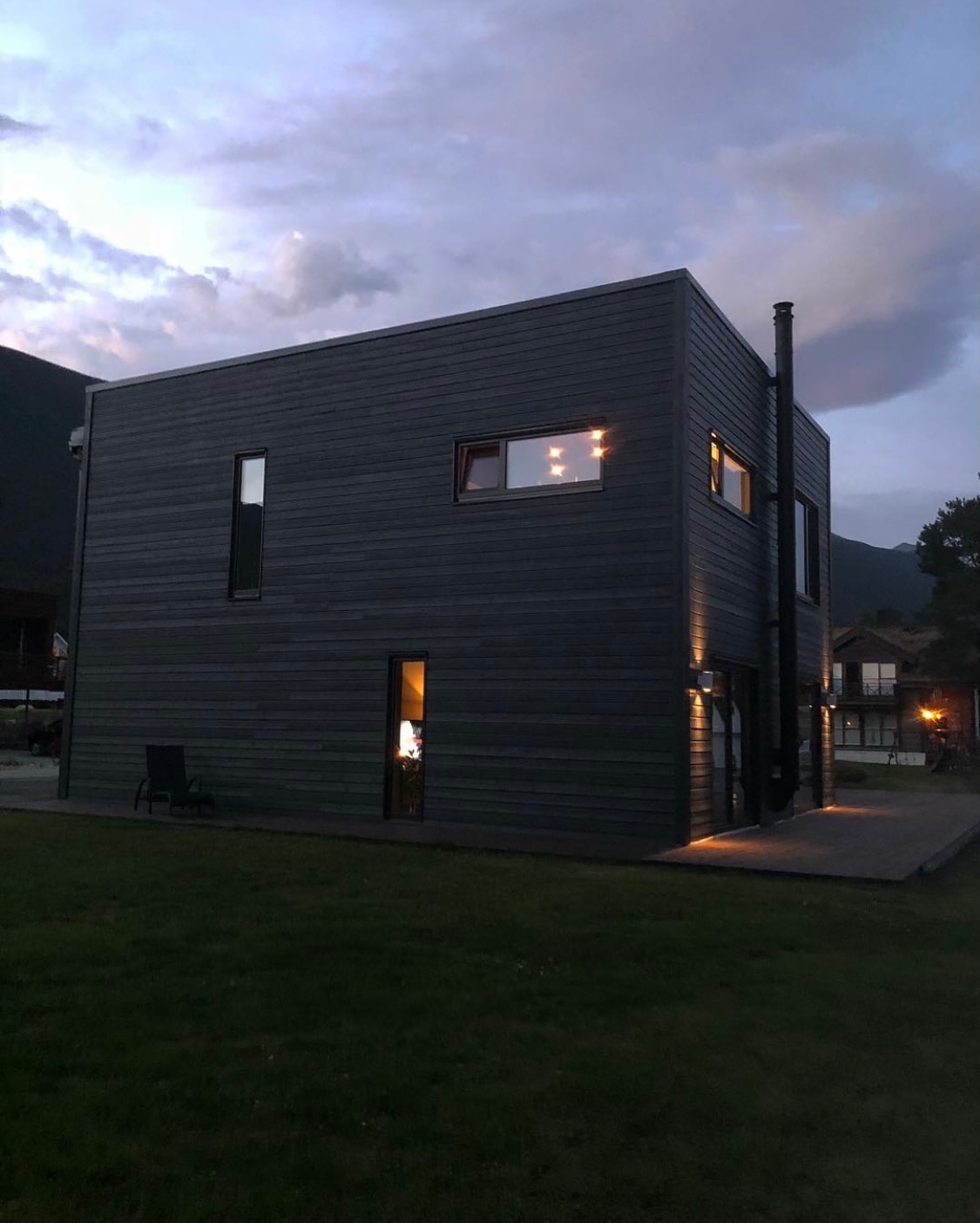 The modern villa is shown at dusk, featuring a sleek wooden exterior with simple lines. Soft lighting emanates from the windows, highlighting the geometric structure. A grassy area surrounds the villa, with a stone patio visible in the foreground.