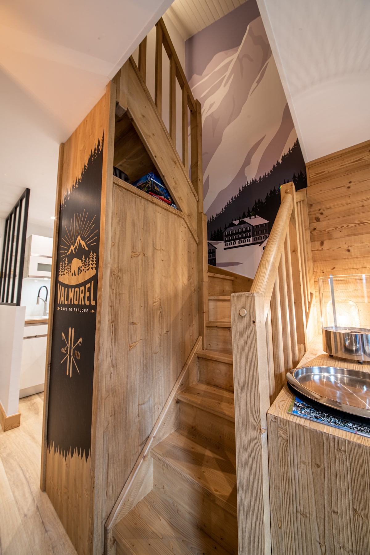A wooden staircase ascends to a second level, surrounded by warm wooden walls that reflect a cozy ambiance. A mural depicting mountains is featured on the wall beside the stairs. A glimpse of the modern kitchen cabinetry can be seen in the background.