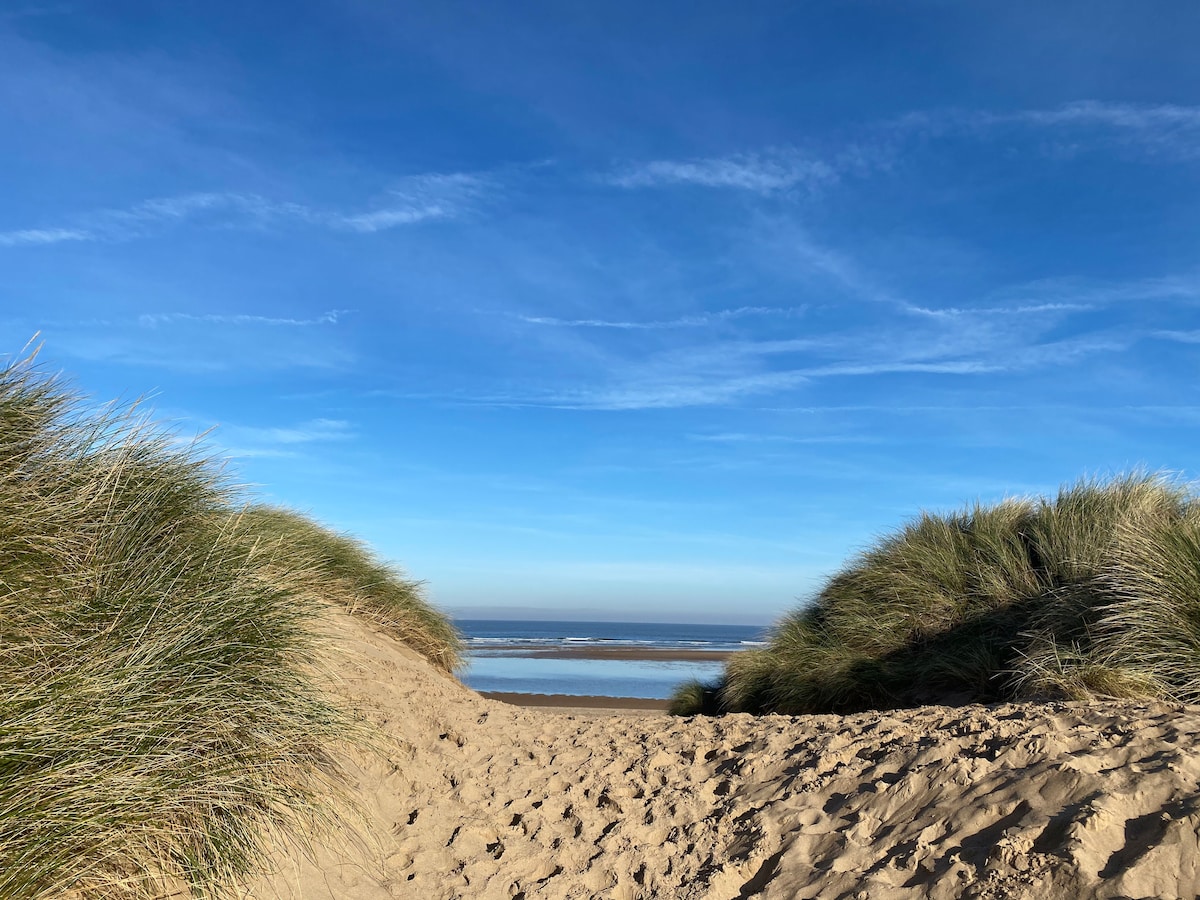 A sandy pathway is flanked by natural grasses leading to a serene beach. The blue sky stretches above, with wispy cloud patterns visible. The gentle waves of the sea are seen in the distance, creating a tranquil coastal scene.