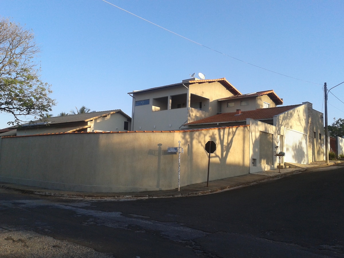 A spacious house is seen from the street corner, featuring a light-colored exterior and a tiled roof. The structure has multiple levels, with several windows and balconies visible. The surrounding area includes a large wall enclosing the property, with trees providing shade in the background.