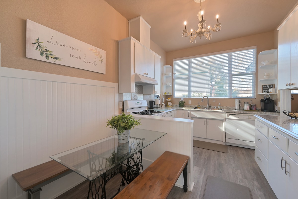 A clean, bright kitchen features white cabinetry and a gas stove with a modern vent hood. Natural light floods through the large window, illuminating a glass-top dining table with a bench and a decorative plant. A chandelier adds subtle elegance to the space.