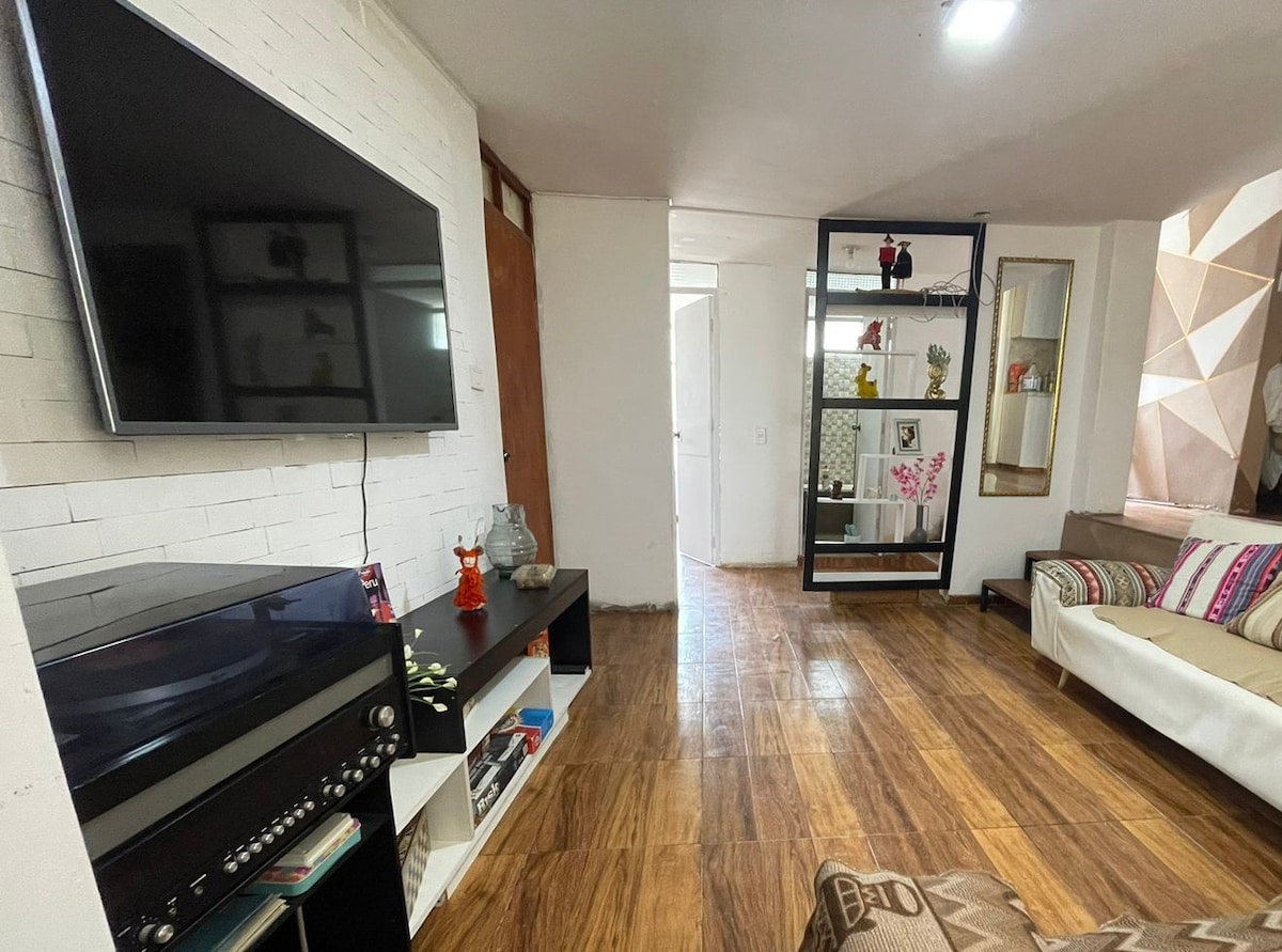 An inviting living area is presented, featuring a flat-screen television mounted on a white-brick wall. A spacious sofa is complemented by colorful cushions. A decorative shelving unit holds various items, while natural light fills the room through a nearby door.