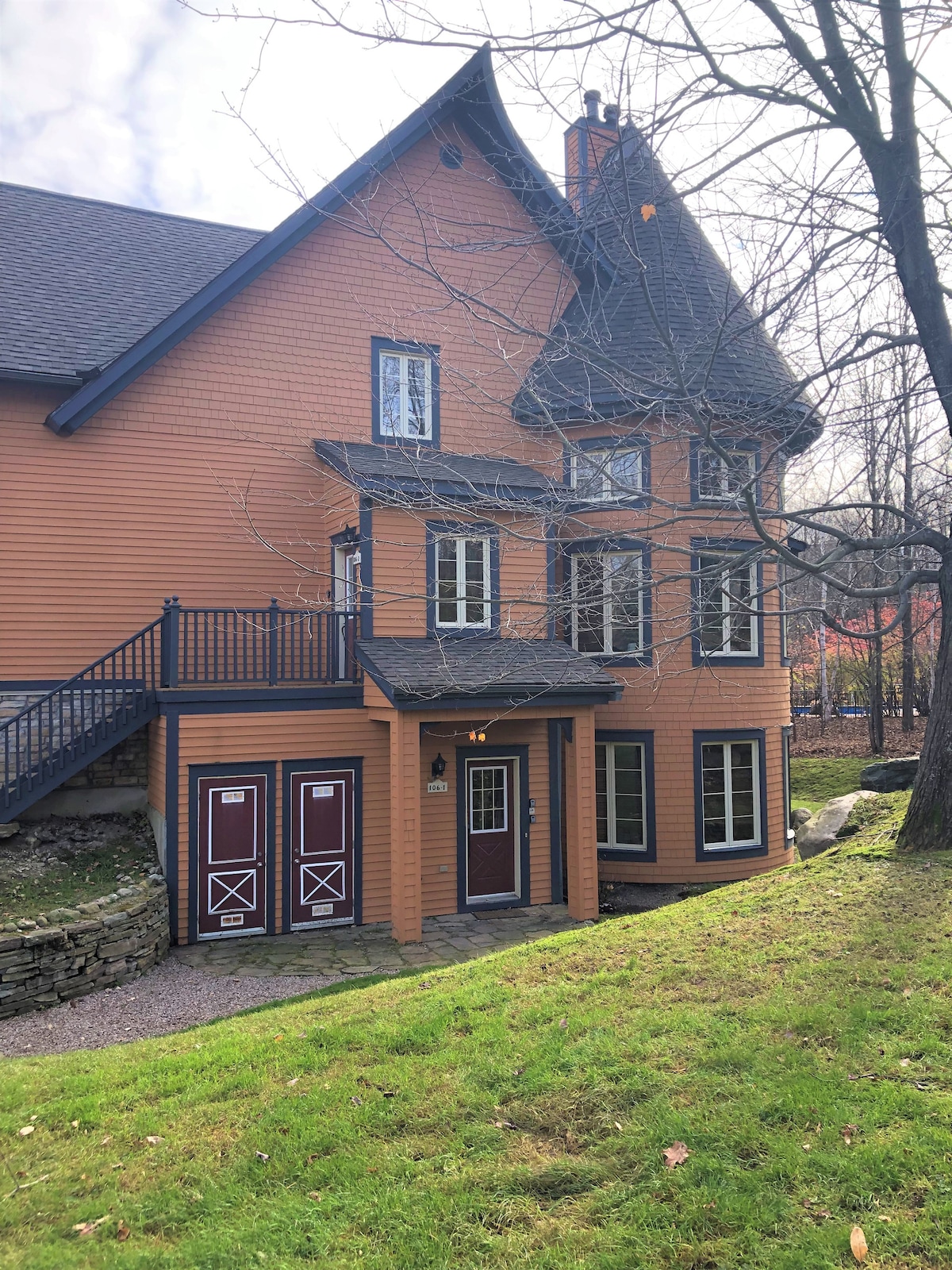 The exterior of a multi-level orange-hued house is framed by lush green grass and bare trees. Two prominent doors, painted in deep red, lead into the building, with multiple windows allowing natural light to fill the interior.
