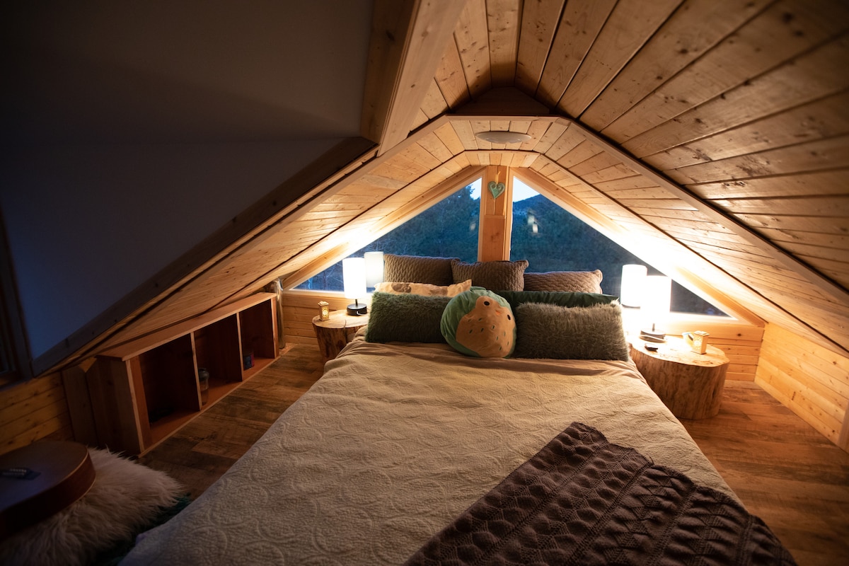 A cozy sleeping loft with a sloped wooden ceiling features a spacious bed adorned with soft linens. Two bedside lamps provide gentle illumination beside the bed, while warm wood accents and a plush rug add comfort. Large windows showcase the surrounding natural scenery.